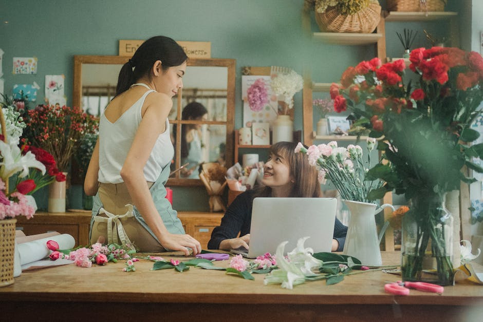 A young female florist with dark hair styled in a ponytail, wearing a white sleeveless top and beige apron, standing at a wooden worktable, conversing with a female customer with shoulder-length brown