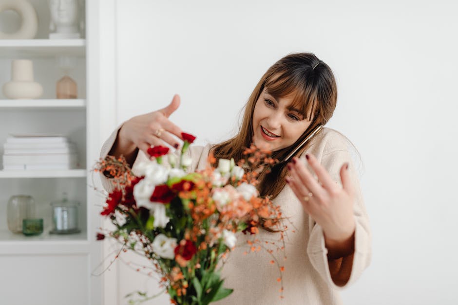 A woman with shoulder-length brown hair, smiling and wearing a beige sweater, is holding a colorful bouquet of fresh flowers while talking on a smartphone. The bouquet includes red, white, and orange 