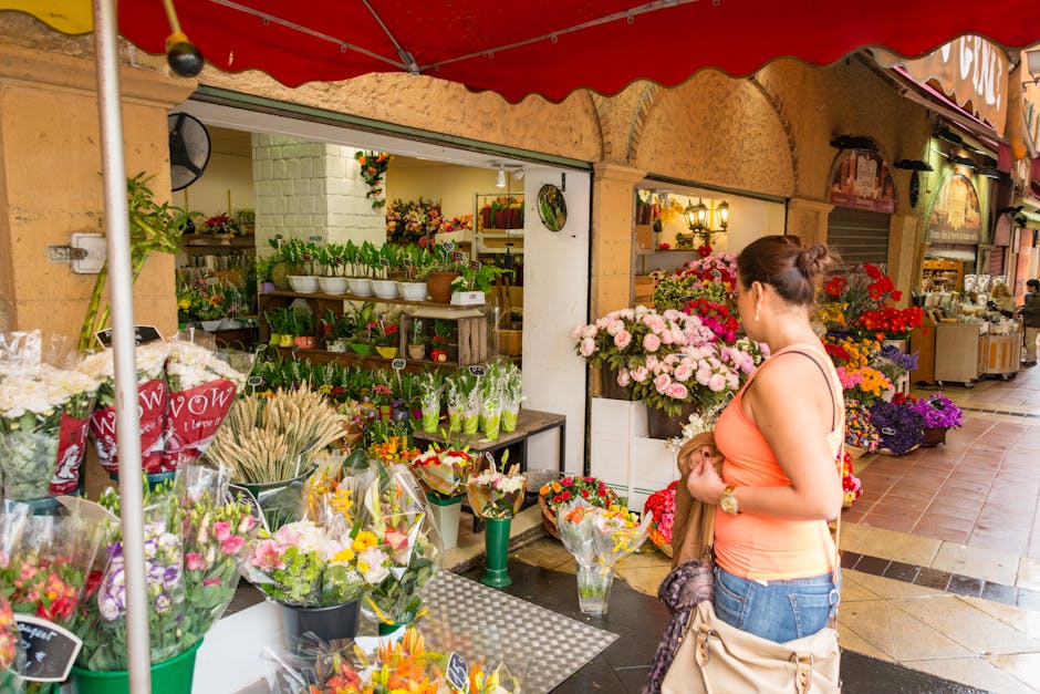 A woman with her hair tied up, wearing an orange sleeveless top and blue jeans, is standing outside a flower shop, holding a bouquet of flowers wrapped in brown paper. The shop displays a vibrant arra
