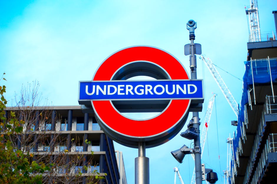 A close-up of the London Underground sign featuring a red circle and blue bar with white 'UNDERGROUND' lettering, mounted on a metal pole against a backdrop of modern multi-storey buildings, some unde