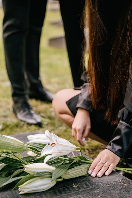 A woman kneeling on the ground arranging a bouquet of white lilies with lush green leaves, creating a floral tribute. She is wearing a black dress with long sleeves, and her hands gently place the flo