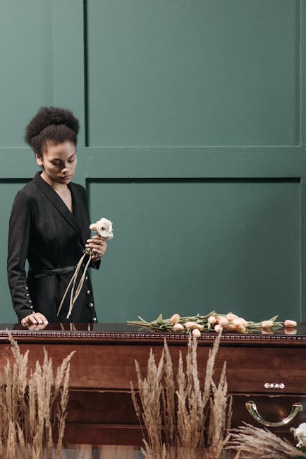 A woman with dark, curly hair styled in an elegant updo, dressed in a sleek black outfit, stands solemnly by a wooden casket with a green backdrop. She holds a small white flower, likely a rose or car